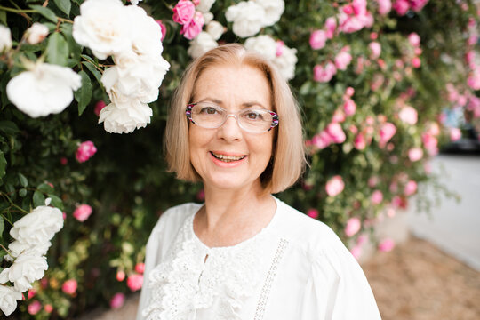Fototapeta Stylish blonde mature woman 55-60 year old wearing vision eye glasses over rose flowers outdoors close up. Looking at camera. Summer season.