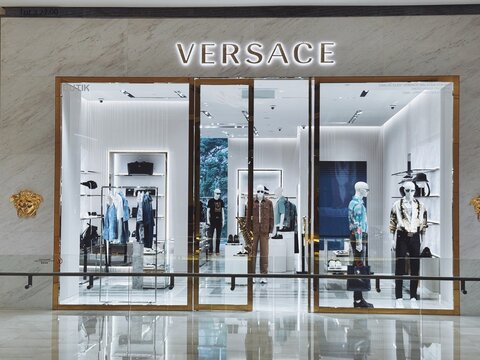 Versace storefront with bold white lighting, gold-framed doors, and mannequins in patterned outfits. Modern luxury fashion display.