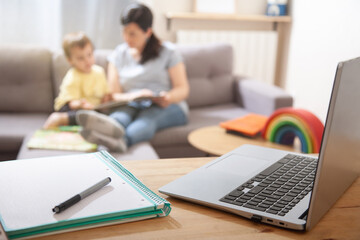 Close-up of home workspace with laptop and notebook, mother and child reading in background