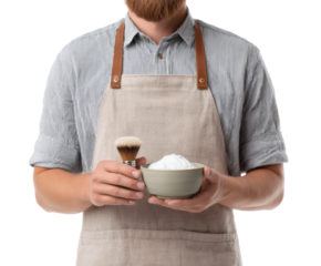A barber, in an apron, is holding a shaving brush and a bowl of shaving cream.