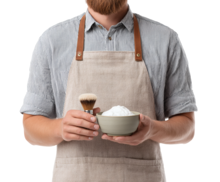 A barber, in an apron, is holding a shaving brush and a bowl of shaving cream.