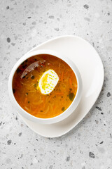 A bowl of golden clear soup with a boiled egg slice, served on a white plate over a textured gray surface. The broth is rich with herbs and vegetables, offering a warm and appetizing presentation