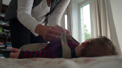 Mother fastening baby’s overalls, adjusting straps while baby remains calm on the bed, nurturing and caring moment in a relaxed home environment during daily routine