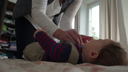 Mother fastening baby’s overalls, close-up hands adjusting straps while baby lies on the bed,...