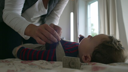 Mother fastening baby’s overalls, close-up hands adjusting straps while baby lies on the bed, calm interaction in cozy room with a mother and baby in morning routine