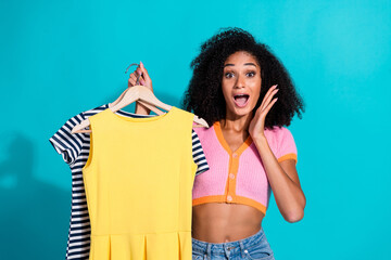 Excited woman holding colorful dresses on hangers against a turquoise background.