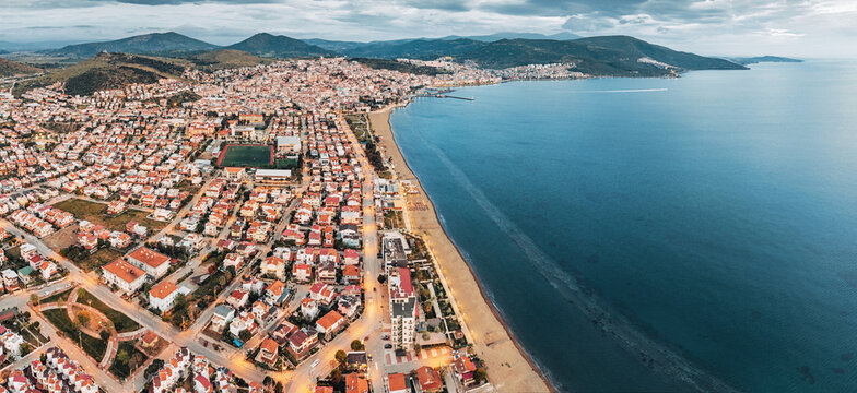 Drone capturing a panoramic view of a Dikili, Turkiye coastal cityscape at twilight, with illuminated streets, residential buildings, and sandy beach