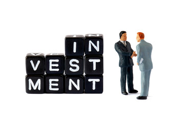 Two miniature businessmen figurines shake hands beside black letter blocks spelling "INVESTMENT" on a white background. This scene symbolizes a business deal or partnership in the context of financial