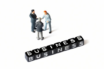 Three miniature businessmen figurines shake hands, with a third standing near them, while black letter blocks spelling "BUSINESS" lie in the foreground. This scene conveys a concept of teamwork and ag