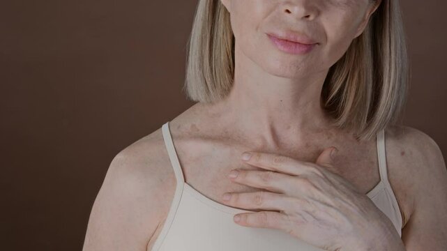 Close up studio shot of senior blonde woman gently touching her shoulder and chest with age spots on naturally aging skin while standing against brown background