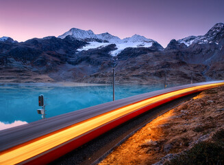 Moving modern red train near azure mountain Bianco lake, snowy rocks and purple sky at night in autumn. Bernina Express, Switzerland. Blurred train in Alps, reflection in water in fall at dusk. Nature
