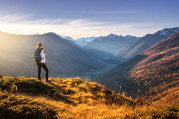 Young woman with backpack standing on grassy mountain ridge with view of valley with high mountain...