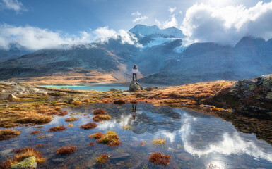 Young woman standing on a rock with stunning view of snowy mountains reflecting in clear alpine lake at sunrise in autumn. Hiking in swiss alps in fall. Girl, pond, orange grass, sky with sun, clouds