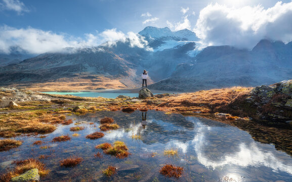 Young woman standing on a rock with stunning view of snowy mountains reflecting in clear alpine lake at sunrise in autumn. Hiking in swiss alps in fall. Girl, pond, orange grass, sky with sun, clouds