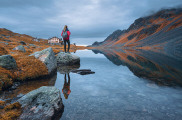 Young woman hiker with backpack on a rock in autumn. Girl and breathtaking view of a mountain alpine lake reflecting the surrounding peaks, orange grass and cloudy dramatic sky. Switzerland in fall
