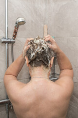 Naklejka premium A view from behind of a person standing in a shower stall. The person is washing his hair, there is foam on his hair. The background is a light tile.