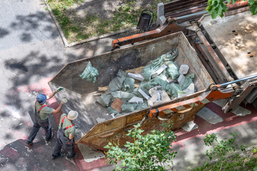 Workers loading construction debris into dumpster on urban street sidewalk.