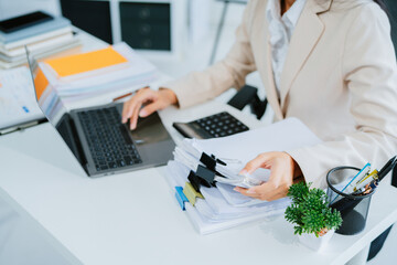 A young Asian businesswoman works alone at a desk with a laptop, stacks of papers and documents. She looks happy and successful managing accounts and preparing financial statements during the day.