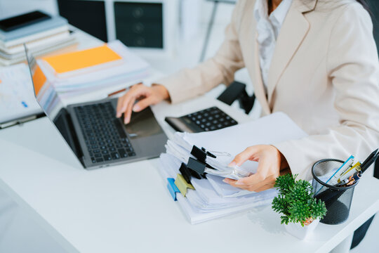 A young Asian businesswoman works alone at a desk with a laptop, stacks of papers and documents. She looks happy and successful managing accounts and preparing financial statements during the day.