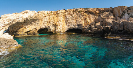 Rugged limestone cliffs with natural sea caves near Ayia Napa, Cyprus, surrounded by turquoise waters revealing a rocky seabed under sunlight.