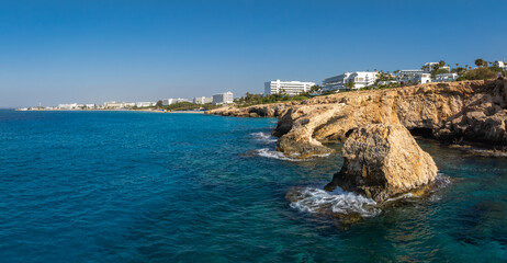 Ayia Napa coastline in Cyprus featuring clear blue waters, golden rock formations, modern white hotels, lush greenery, and palm trees.