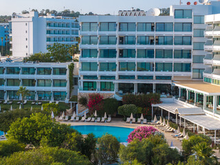 A modern multi story hotel with glass windows and balconies, a central pool with sun loungers,...