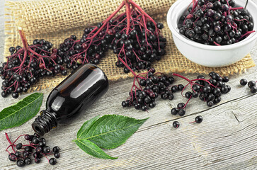 Delicious homemade black elderberry syrup in glass jar and bunches of black elderberry with green leaves on wooden desk. Sambucus.