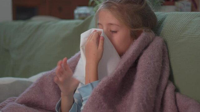 Sick girl blowing nose into tissue while sitting on sofa at home, looking tired and unwell with cold and flu symptoms at night