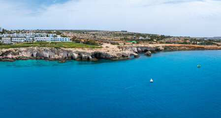 Fototapeta premium Rugged limestone cliffs with Love arch caves along the Ayia Napa shoreline, turquoise Mediterranean waters, and modern houses in a rocky, sparse landscape.