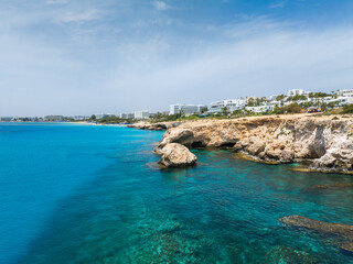 Fototapeta premium Turquoise waters and rugged rock formations in Ayia Napa, Cyprus, with modern white luxury hotels and resorts along the shoreline under a clear sky.