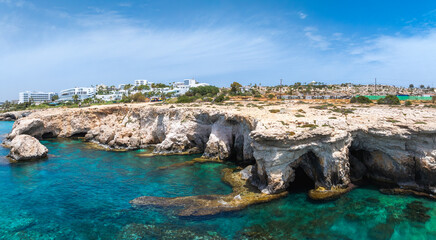 Rugged limestone cliffs with sea caves meet turquoise waters in Ayia Napa, Cyprus. White luxury resorts and greenery are visible in the background.