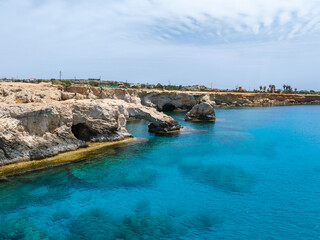 Turquoise waters meet rugged limestone cliffs in Ayia Napa, Cyprus. The shoreline features rock formations Love arch, caves, sparse vegetation, and a bright blue sky.