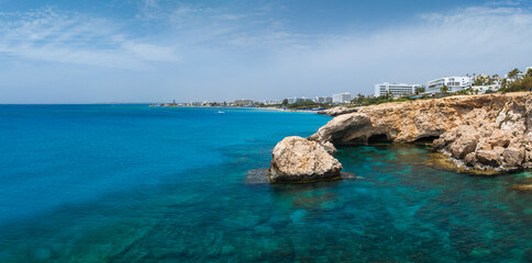 Turquoise waters and rugged rock formations in Ayia Napa, Cyprus, with modern white luxury hotels and resorts along the shoreline under a clear sky.