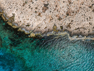 Aerial view of a rocky coastline in Ayia Napa, Cyprus, with jagged edges, scattered vegetation, and vibrant turquoise waters of the Mediterranean Sea.