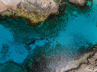 Aerial view of a rocky coastline in Ayia Napa, Cyprus, with clear turquoise waters, rugged terrain, and visible underwater textures and gradients.