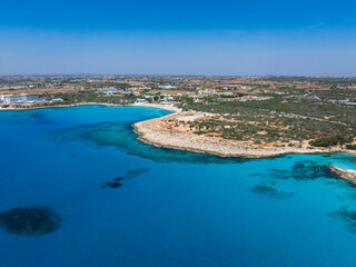 Aerial view of Ayia Napa, Cyprus, showing turquoise waters, rocky shores, scattered buildings, resorts, lush greenery, and a clear blue sky.