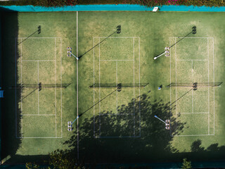 Aerial view of a sports field featuring multiple marked courts, tall light poles casting shadows, and a tree shadow partially covering the corner.