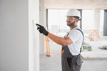 Indian Male builder in work overalls plastering a wall using a construction trowel.