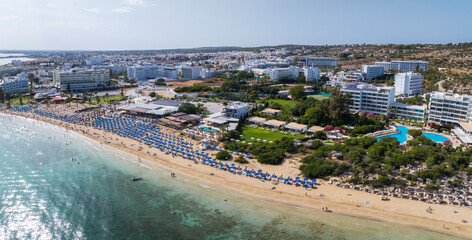 Aerial view of Ayia Napa, Cyprus, featuring sandy beaches, turquoise waters, blue umbrellas, sunbathers, luxury hotels, and lush greenery.