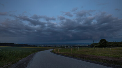Landschaftsaufnahme mit Regenwolken, Feldern, Wiesen, Bäumen und Strasse.