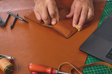 Leather craftsman gluing a brown leather wallet in his workshop, surrounded by tools and a laptop