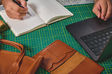 Leather craftsman writing notes in notebook and using laptop while working on leather goods at...