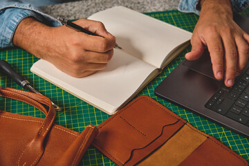 Craftsman working with leather goods using laptop and taking notes in notebook