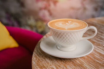 Elegant cup of latte art on wooden table in cozy cafe setting with blurred floral background and colorful cushion