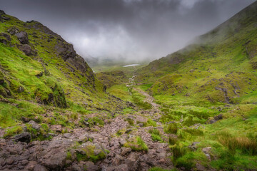 Discover a tranquil Carrauntoohil mountain and valley adorned with lush green slopes and rugged, rocky terrain, all surrounded by a soft mist and a profound sense of serenity, Kerry, Ireland