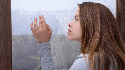 Lonely young lady looking out window, lost in thought, touching glass with rain drops. Depressed...