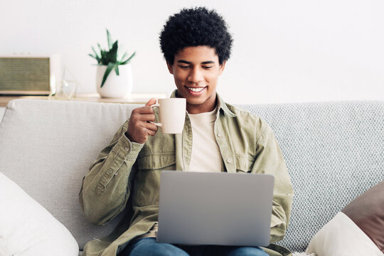 Fototapeta E-learning concept. Handsome black male student using laptop computer and drinking hot coffee at home, panorama. Happy teenage guy listening to online lecture, communicating to tutor on webcam