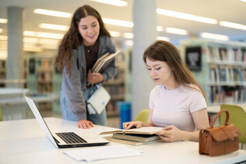 Female student works on laptop in library and talks with female friend. Studying, using additional online sources of information, searching for information on Internet...........