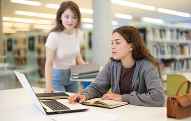 Young female student asks young female student working on laptop in library