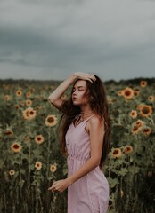 Vertical photo. Woman in pink knee-length dress stands among tall sunflowers. Right hand touches hair. Sunflowers have dark centers and yellow petals. Background shows more sunflowers and cloudy sky.